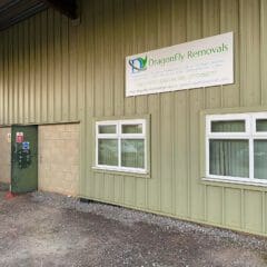 Industrial building exterior with a green metal façade and a small sign reading "Dragonfly Removals" above white-framed windows. A metal door with a warning sign is on the left. Gravel pathway in front.