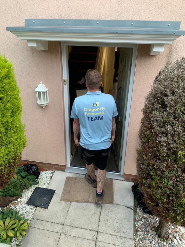 A person wearing a light blue "DragonFly Removals Team" shirt is entering a house. They have tattoos on their arms and are wearing dark shorts and sneakers. The house has light pink walls and is surrounded by trimmed shrubs.
