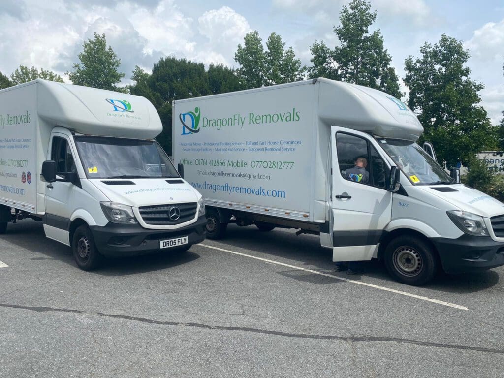 Two white "Dragonfly Removals" trucks parked side by side in a parking lot. They have green and blue logos, and contact details printed on the sides. The sky is partly cloudy, and there are trees in the background.
