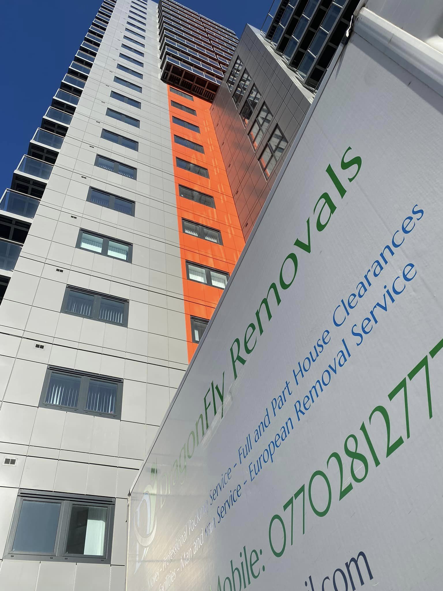 High-rise building with orange and white sections against a blue sky. In the foreground, a moving van is partially visible, displaying "DragonFly Removals" with contact details and services offered.