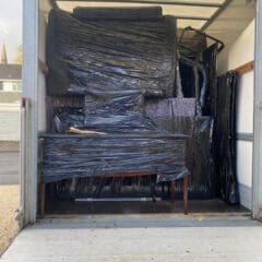 A moving truck loaded with furniture wrapped in black plastic for protection. The items are tightly packed and stacked inside the vehicle, including a large table with a wooden handle poking out in the center. The truck is parked outdoors.
