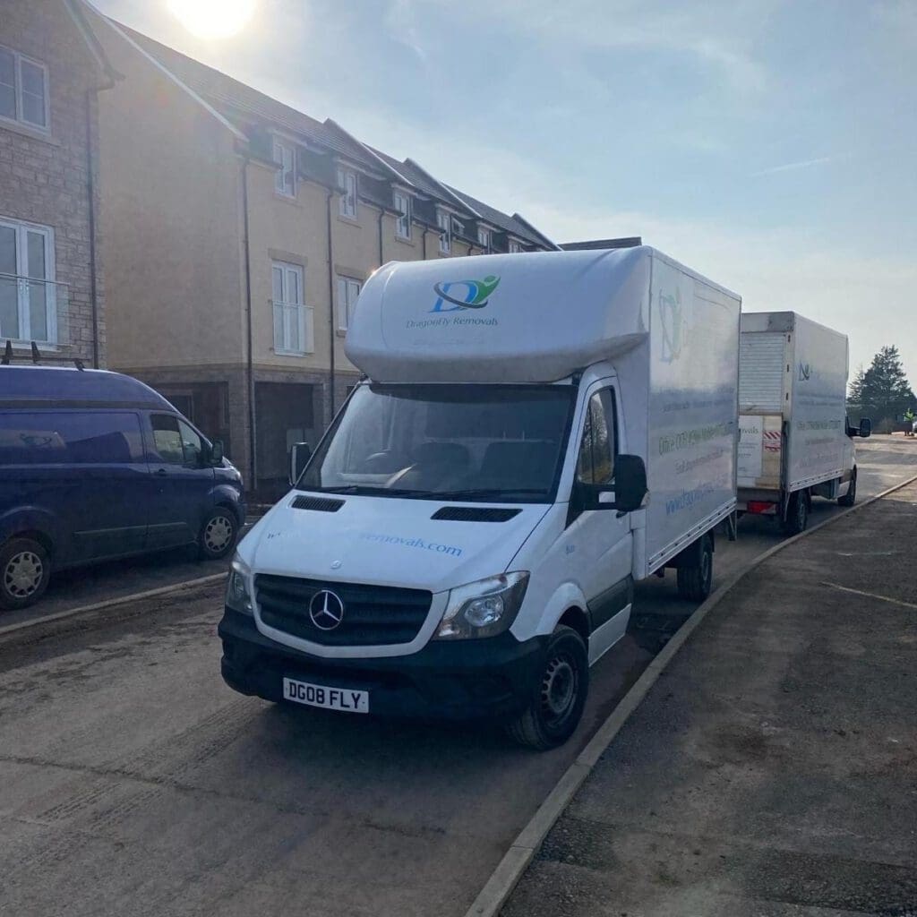 A white delivery truck with a Mercedes logo is parked on a residential street in front of townhouses. The truck has the logo and name "Door2Door" printed on it, along with a website address. A blue van is parked nearby. The sky is clear and sunny.