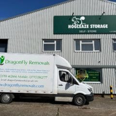 A white moving truck with "DragonFly Removals" branding is parked in front of a corrugated metal building labeled "Hogleaze Storage Self-Store." The truck displays contact information for the removals company.