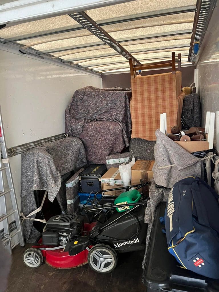 A moving truck filled with furniture and boxes. Items include a lawnmower, stacked chairs, and covered furniture. Blankets cover much of the load for protection. A ladder leans against the side.