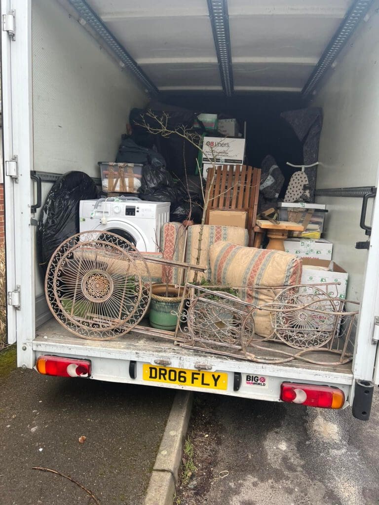 An open truck is filled with various items, including boxes, a washing machine, furniture, and a decorative metal bicycle. The truck's open back door shows the packed contents. The license plate reads "DRO6 FLY.