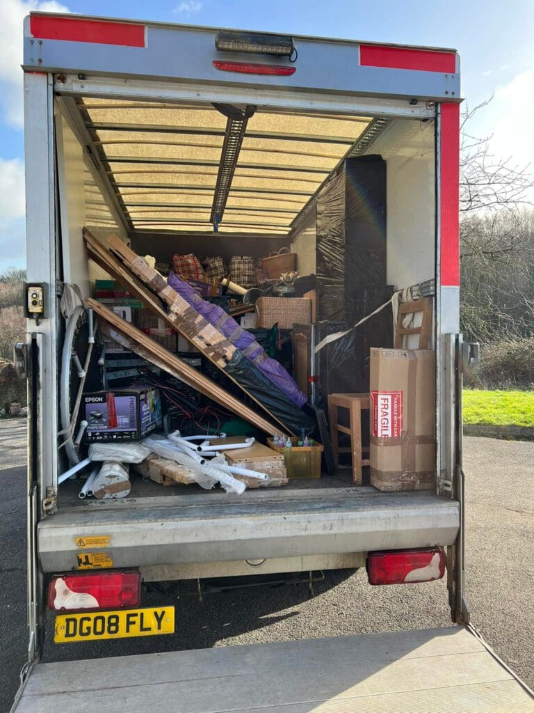 Open moving truck filled with various household items, including furniture, boxes, and a rolled rug. The truck is parked on a paved surface, with greenery and trees visible in the background under a partly cloudy sky.
