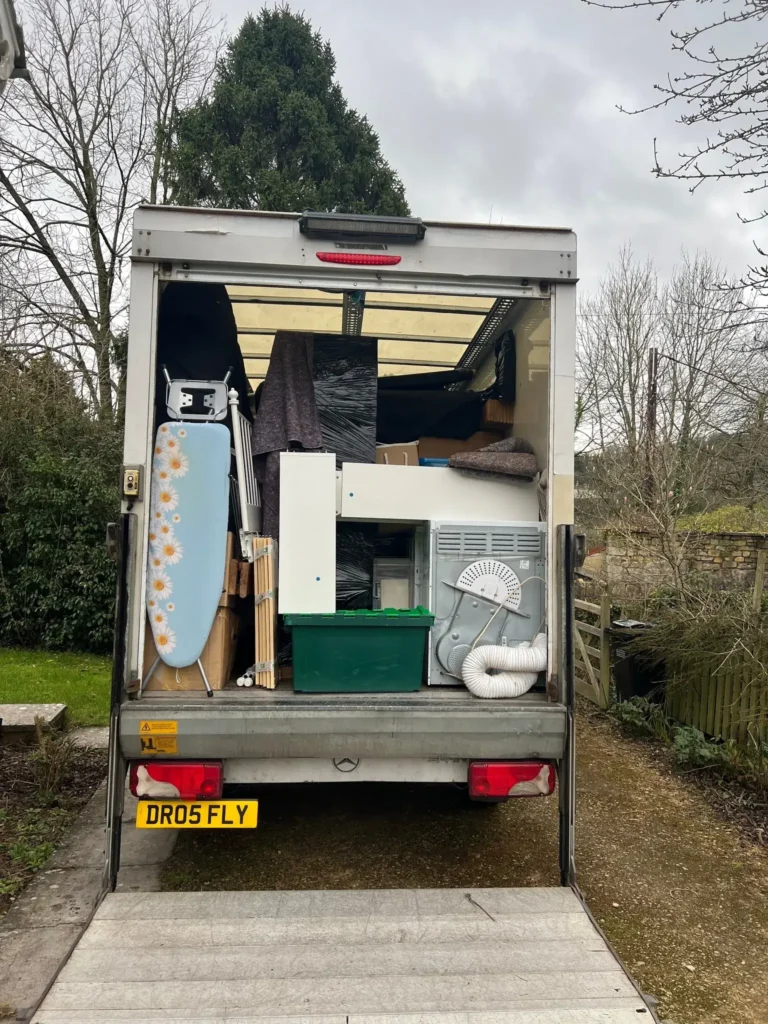 A moving truck is parked with its back open, revealing various household items packed inside, including an ironing board, a cabinet, a green storage bin, and an air conditioning unit. Trees and a cloudy sky are visible in the background.
